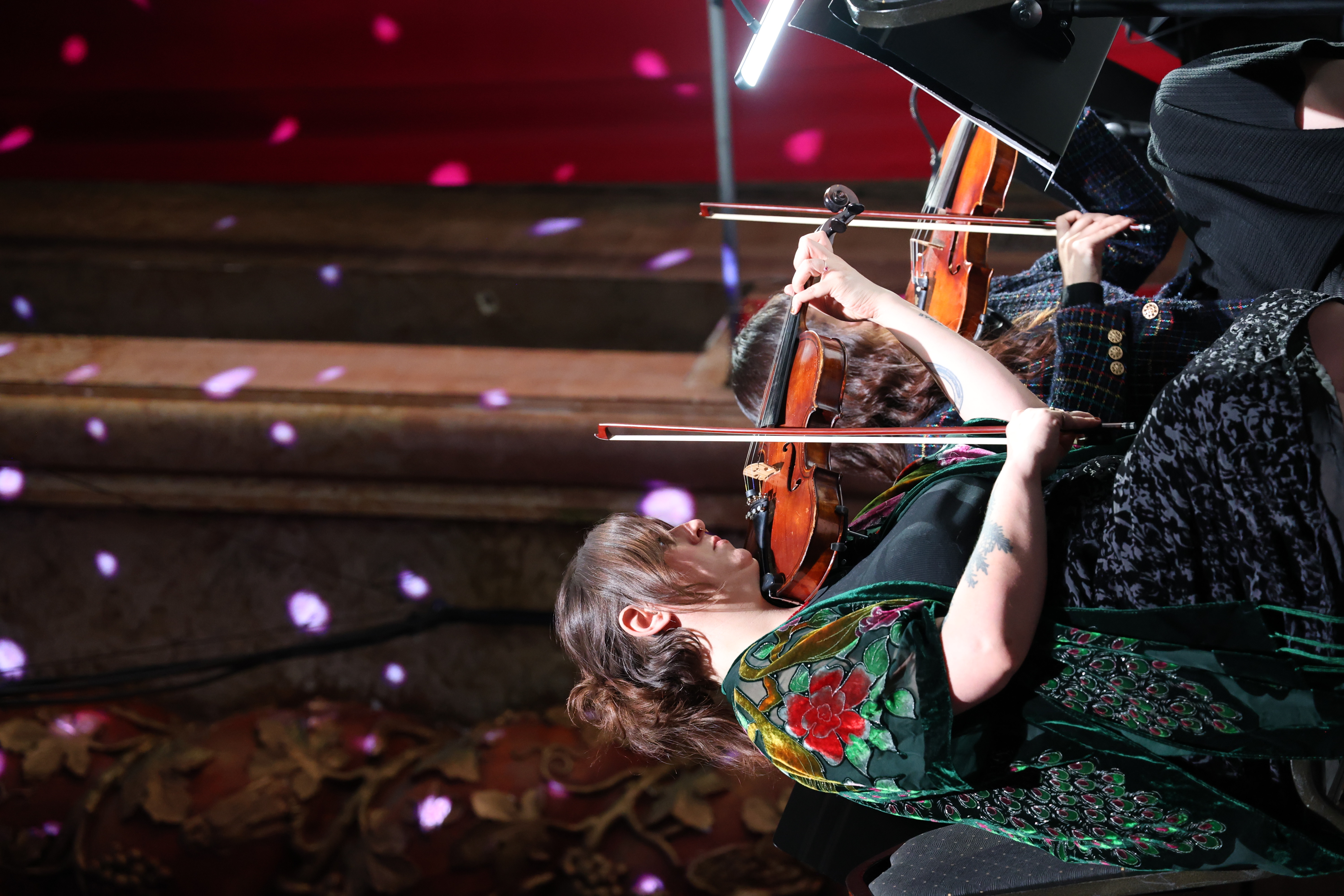 Violinist playing on stage, floral shawl, and dark background.